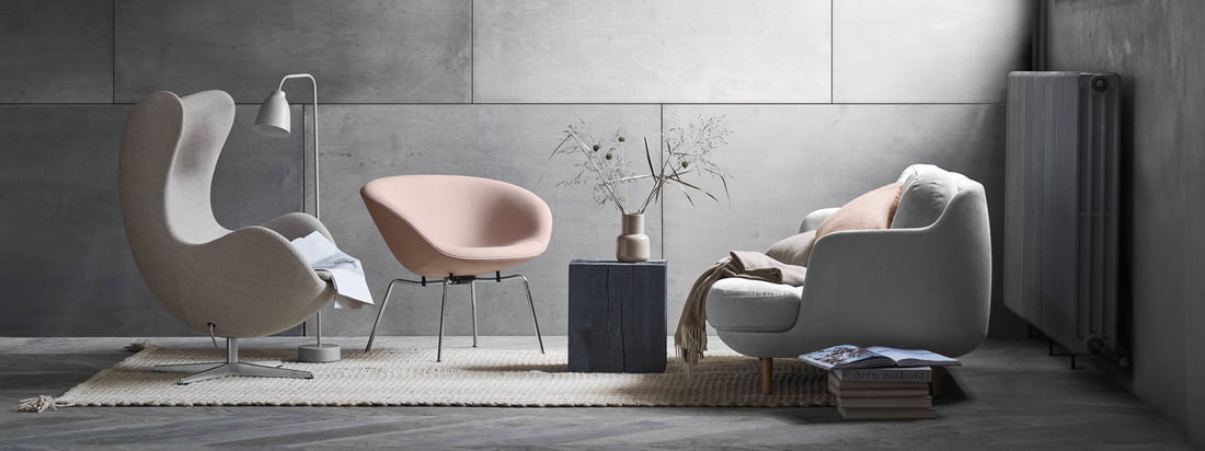 Le confortable canapé 2 places Lune de Fritz Hansen avec le fauteuil pot assorti dans des tons doux de gris et de rose. Une ambiance de salon moderne devant un mur gris.