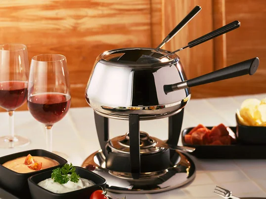 Le service à fondue Classic de l’entreprise suisse Spring apparaît dans la forme traditionnelle légèrement bombée avec un long manche sur un réchaud esthétique.