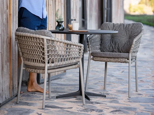 Ocean Chaise avec accoudoirs, taupe de Cane-line dans la vue d'ambiance. Un dossier et des accoudoirs bombés offrent un confort agréable, ce qui permet de passer des heures agréables dans cette chaise.