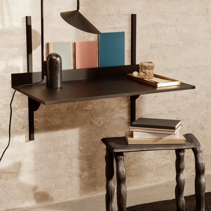 Le bureau Sector de Ferm Living accroché au mur en combinaison avec des livres, une lampe à poser et un tabouret.